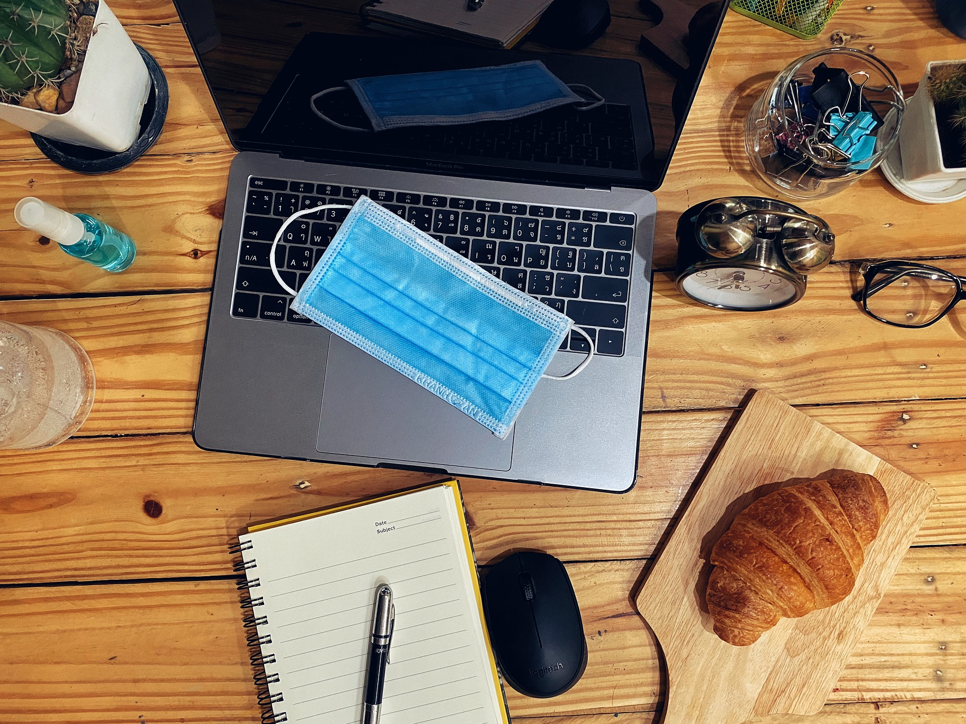 A workspace with a laptop, notebook, pen, mouse, croissant, eyeglasses, alarm clock, and a face mask on a wooden desk.