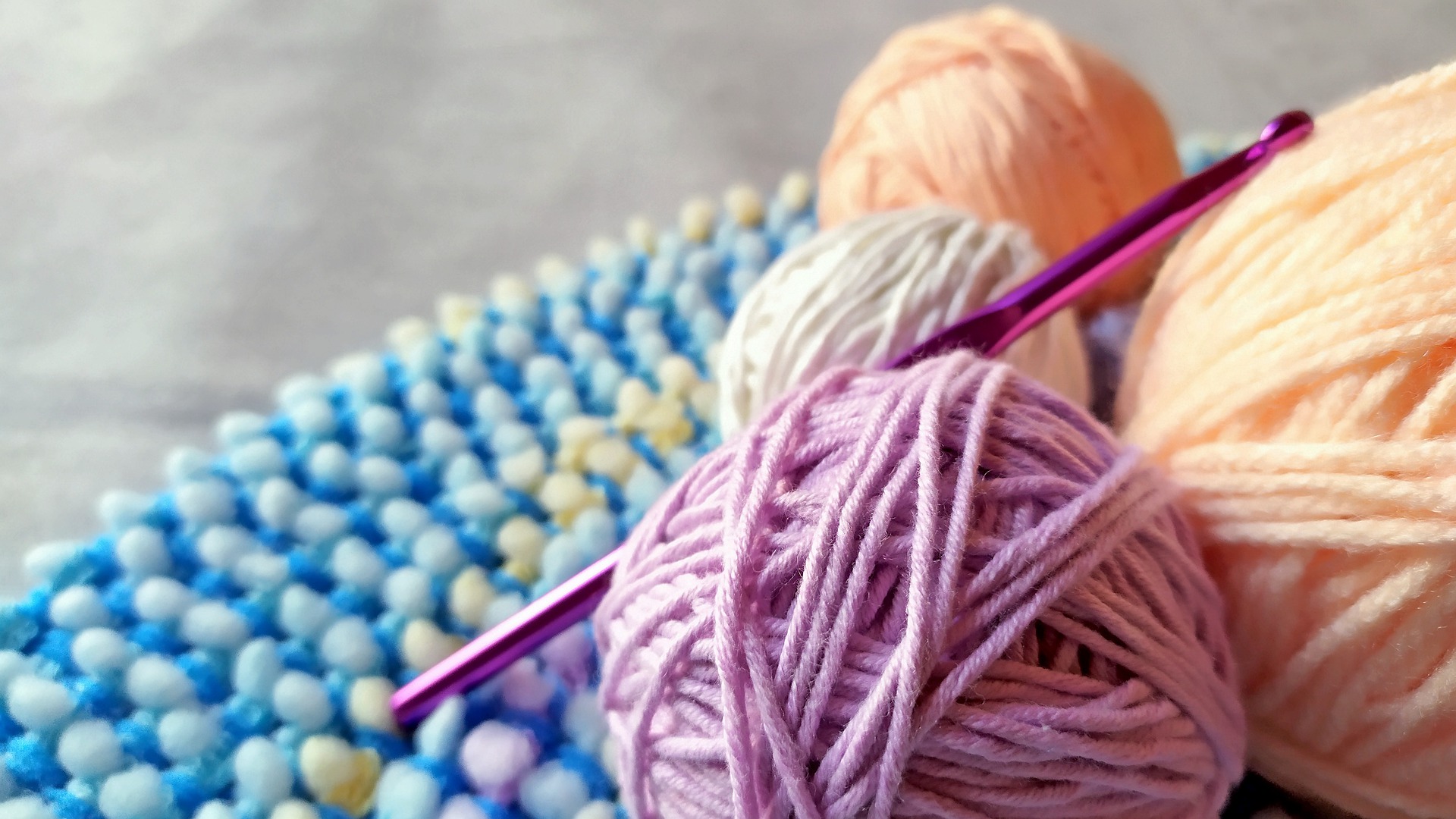 A a close-up of colorful yarn balls and a purple crochet hook. They are all resting on a blue crochet project.