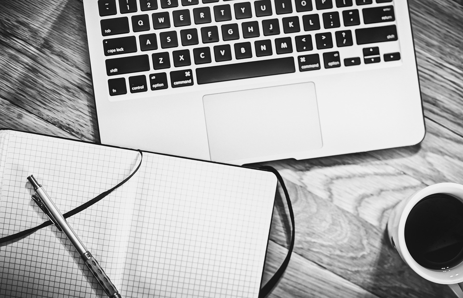 A black and white image of a laptop, a notebook, a pen, and a cup of coffee on a wooden table, suggesting a productive workspace.