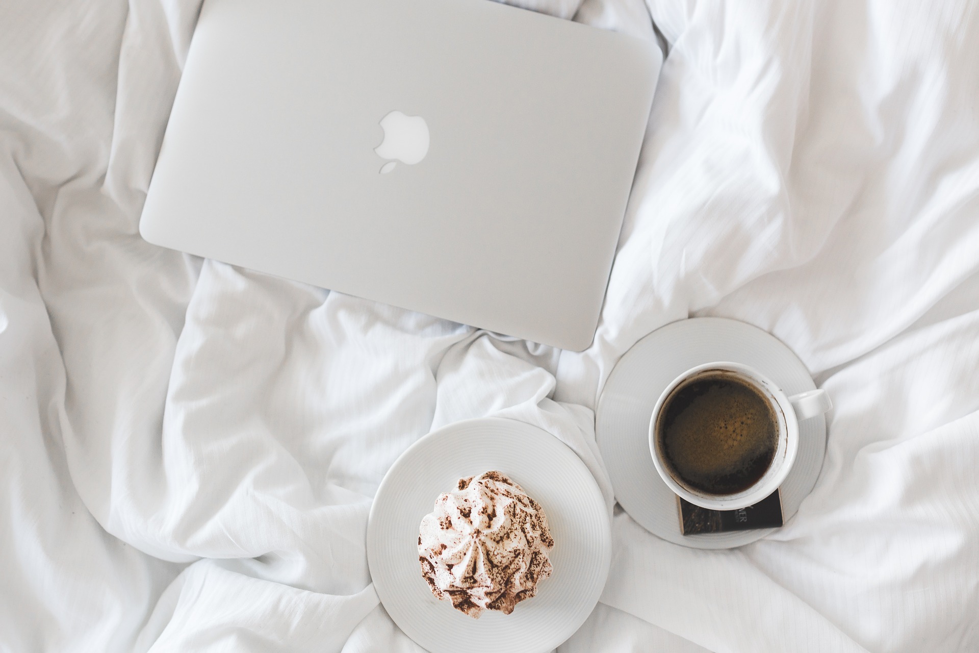 A laptop, a cup of coffee, and a pastry on a white bed. It suggests a cozy and productive workspace in a bedroom.